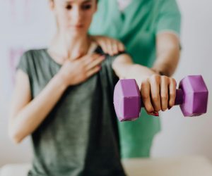 Crop unrecognizable orthopedist in uniform helping slender female in casual wear reaching arm with dumbbell in doctor office in hospital